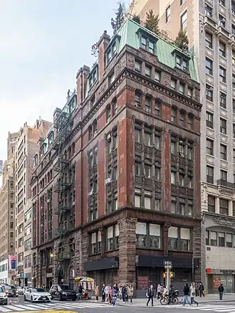 A dark red brick apartment building, with brown stone facing the lower two of its seven stories, topped by a green mansard roof with dormer windows, seen from the opposite corner of its intersection. Taller buildings are beyond it, and at street level is a storefront with "Estex" written above it.