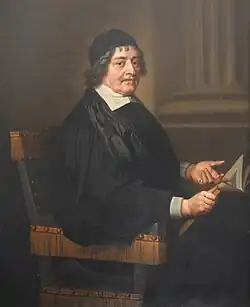 A man with a black cap and gown, seated. He is holding an open book in his right hand and pointing to it with his left