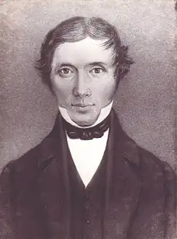 A young clean-shaven man with wavy hair, wearing dark coloured jacket and vest over a white shirt with a high collar. He is looking straight ahead, with a faint smile