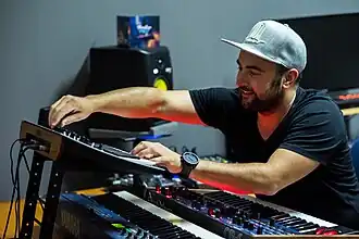 Jordy Leenaerts wearing a black t-shirt and light grey cap, playing a set of stacked keyboards, grinning