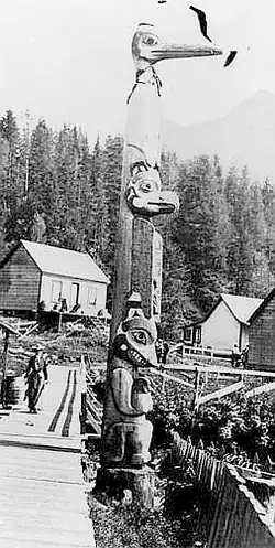 Black-and-white photograph of a Tlingit totem pole in Ketchikan, Alaska, circa 1901, carved with stacked human and animal figures, with wooden buildings and forest in the background.
