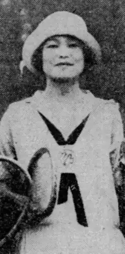 A smiling young Japanese woman wearing tennis whites, wearing a hat, holding a racket