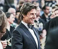 Tom Cruise, wearing a tuxedo, is smiling while standing among a group of people