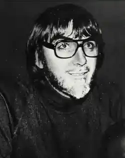 A monochrome photo of a short-bearded man with medium-length hair, wearing glasses with thick black rims.