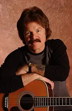Johnston playing a Paul Reed Smith Artist's Gold Top while wearing a black t-shirt and jeans, in front of colorful stage lights
