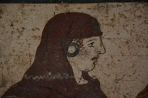 Up close of one of the dancing women, she has a reddish brown head covering, large black circular earrings, red curly hair peaking out around her face, a angular nose and a very solemn look on her face as they all dance.