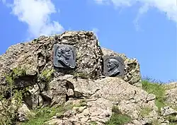 Two plaques as momuments on a cliff wall, a woman's face on the left facing right in the foreground, and a man's face on the right facing left slightly behind