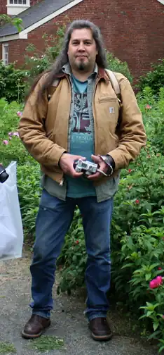 Photograph of Tommy Joseph, a Tlingit woodcarver and sculptor from Sitka, Alaska, standing on a garden path and holding a camera, with greenery and a brick building behind him.