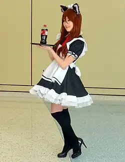 A young woman wearing a maid outfit with cat ears, presenting a bottle of soda on a tray.