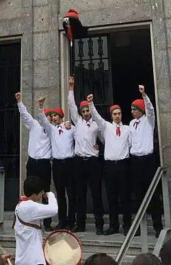 Five young men, arms raised, wearing white shirts, black pants and orange caps