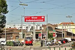 A red historic tram leaving a large brick tram depot, now the Vienna Transport Museum, with a large red sign reading "Wiener Straßenbahnmuseum"