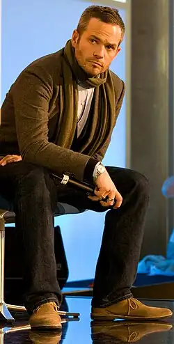 A man dressed in a blazer, scarf, and jeans, sits on a stool while holding a microphone