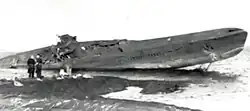 A damaged submarine on a sandy beach with two people standing nearby.
