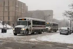 Two University of Chicago shuttle buses (with "UGo" red branding on the sides) on 57th Street outside the Regenstein Library during a snowstorm