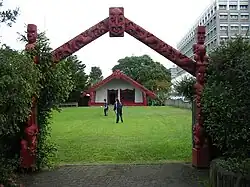 A Māori carved gate way frames a Māori meeting house with two active young people in between on the grass area.