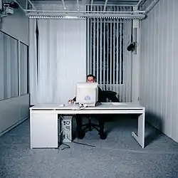 A man seated at a computer desk in a carpeted white room, with a window.