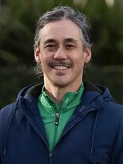 A middle-aged man with mid-length greying hair and who is wearing a green and blue jacket smiles at the camera