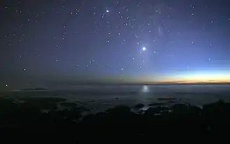 A photograph of the night sky taken from the seashore. Many glimmers of sunlight is on the horizon. There are many stars visible. Venus is at the center, much brighter than any of the stars, and its light can be seen reflected in the ocean.