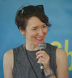 Unposed bust photo of a white woman holding a microphone and looking to the camera's right; she has dark brown hair, a sleeveless grey shirt, and is smiling.