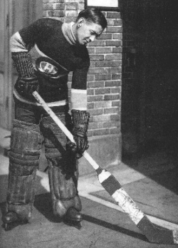 Profile of a goaltender in full uniform looking down at his stick. He is wearing thick pads around his legs, and padded gloves that reach near his elbows.