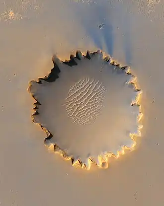 Image 3 Victoria Crater, Mars Photo credit: Mars Reconnaissance Orbiter Victoria Crater, an impact crater at Meridiani Planum, near the equator of Mars. The crater is approximately 800 meters (half a mile) in diameter. It has a distinctive scalloped shape to its rim, caused by erosion and downhill movement of crater wall material. Layered sedimentary rocks are exposed along the inner wall of the crater, and boulders that have fallen from the crater wall are visible on the crater floor. The floor of the crater is occupied by a striking field of sand dunes. The Mars rover Opportunity can be seen in this image, at roughly the "ten o'clock" position along the rim of the crater. More selected pictures