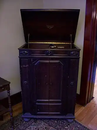 A wooden standing cabinet phonograph record player, about waist-high, photographed from the front, in the corner of a room next to a doorway. The rear-hinged lid at top of the cabinent is opened upwards, held open by a pair of support arms. The record turntable is partly visible peeking above the lip of the cabinet front. A pair of swing doors occupies most of the front of the cabinet.
