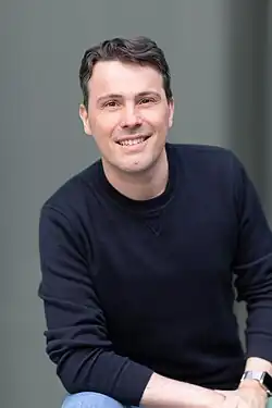 Vincent Pilette looking toward the camera wearing a dark color long-sleeve shirt. A wristwatch and blue jeans are visible