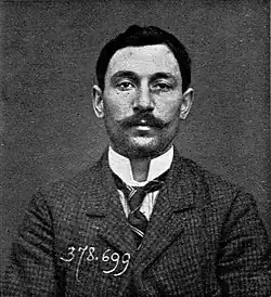 A black-and-white antique mugshot of a man with a mustache wearing a suit and tie, with the number 378.699 written over the image