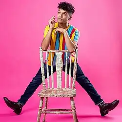 Thomas leans on a chair with his legs splayed, balancing on his heels. He has thick curly hair, light brown skin, and is wearing a striped sweater.