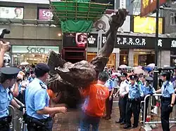 huge bronze sculpture being taken away by removal men dressed in orange, surrounded by police officers in blue shirts and caps or berets and overlooked by crowds