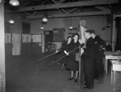A wargame in progress. The men on the far-right command the escort ships and may only view the game board through holes in the screen. The woman in the foreground is Jean Laidlaw.