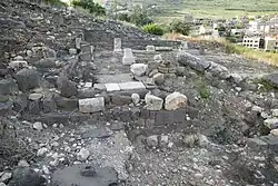 Ruins of the basalt-built synagogue at Khirbet Wadi Hammam on a sloping hillside, with columns and wall stubs in the foreground and modern houses of Wadi al-Hammam visible across the valley behind