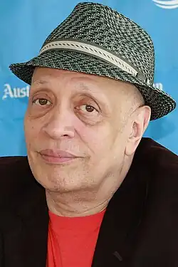 Head and shoulders of Walter Mosley with drooping eyelids wearing black fedora, red shirt without a collar, black jacket, and clean shaven.