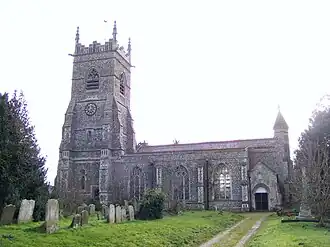 Flint-built parish church at Wangford with a tall square tower featuring a clock face and decorative battlements. The church has large Gothic-style windows along the nave, a tiled roof, and a small entrance porch at the right. Gravestones stand in the grassy churchyard, with a dirt path leading toward the open doorway.