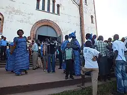 A crowd of people in blue interact on the steps in front of the church entrance. The tall church’s exterior is white, with brown embellishments and a gray-blue door.