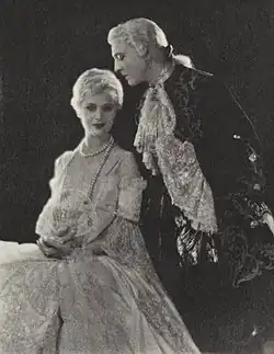 Black and white photograph. Manon, seated, gives the camera a direct and provocative look; des Grieux, eyes closed, leans in as if smelling her hair. Both wear eighteenth-century garb.