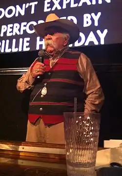 An elderly white male with a long, white moustache. He is wearing a cowboy hat and striped waistcoat while holding a microphone. He is standing in front of a screen.