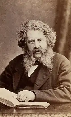 A man with a beard and curly hair sitting at a table with an open book.