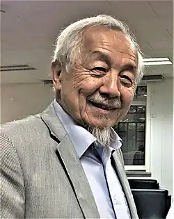 An elderly Asian man with a thin goatee wearing a blue dress shirt and a gray striped sportcoat