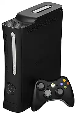 Close up photograph of a black video game console facing to the left as it stands upright on its side. The front has a silver disc tray cover and recessed section with a concave button. A black wireless video game controller sits next to the console.