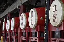 A horizontal view of five large ceremonial drums displayed in a covered gallery at Xi'an's Drum Tower. Each drum features a crimson lacquered barrel body with brass ring hardware, topped with a circular white drumhead inscribed with Chinese calligraphic characters in dark red. The drums are mounted on matching red wooden stands with ornate railings. Dark wooden pillars support the structure above, and a vertical black banner with gold Chinese characters hangs on the right side. Small informational placards are visible at the base of the drums, with one readable text mentioning 'Beginning of Spring'