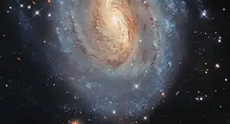 An oval-shaped spiral galaxy, of which only the centre and lower half is in frame. Its centre is mainly golden in colour with a white glowing core, while its thick spiral arms are mostly blue, particularly at the outskirts; the colours merge in between. Dark lanes of dust swirl through the centre, blocking some of its light. Stars and distant galaxies can be seen around the edges on a black background.