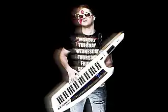 Stylized image of Yinon Yahel with black background, wearing a black t-shirt with white lettering, ripped blue jeans, and white plastic-frame sunglasses, playing a portable keyboard strapped over his shoulder