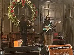 Two performers on a stage. The one on the left is bowing a viola and the one on the right is strumming an electric guitar and singing