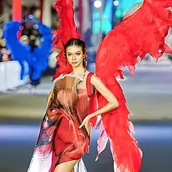 Kato wearing a fanciful dress on a fashion runway, left arm akimbo, looking slightly left of camera
