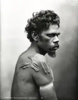Portrait photograph of a shirtless Aboriginal man with a band on his arm and a scarred shoulder, turned away from the camera