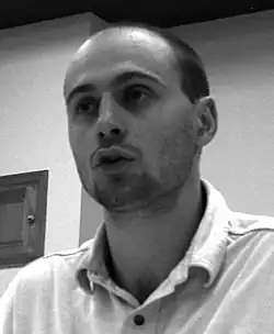 black-and-white image of Yves Engler wearing a light shirt, looking right of camera and appearing to speak