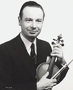 Black and white, dramatic-pose publicity shot, of a clean-shaven man with designer-stubble moustache, big dark eyes, and receding, oiled black hair.