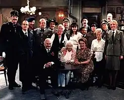 A group photo of eighteen actors in costume for the sitcom 'Allo 'Allo!, posed inside a pub. The cast includes actors dressed in army uniforms, waitresses, and various civilian outfits