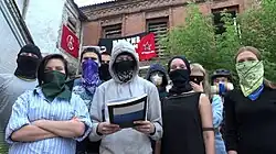 People wearing balaclavas and bandanas standing in formation in front of an old brick building, which has red banners on it